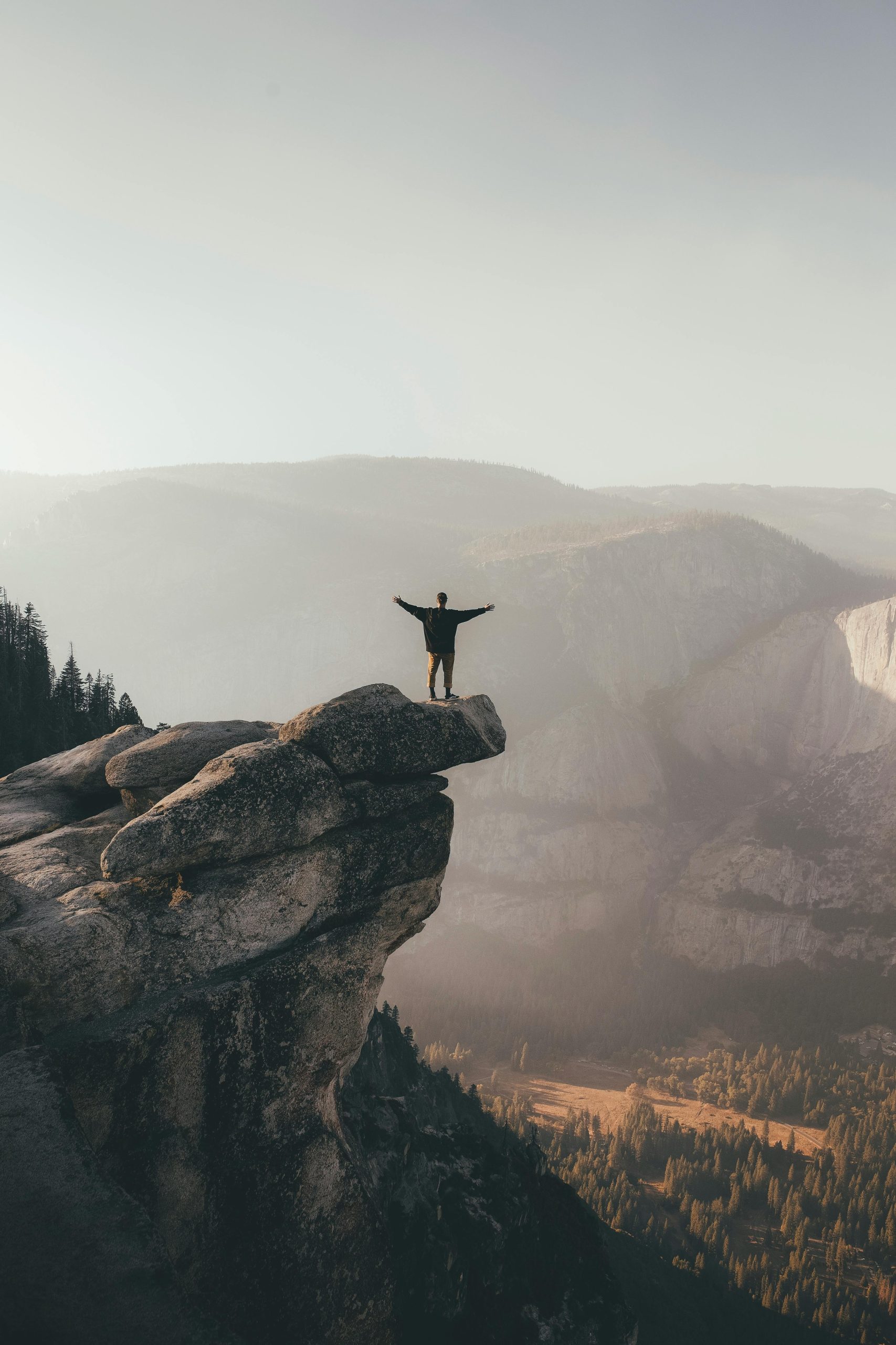 Person with arms raised standing on cliff edge, surrounded by scenic mountain landscape, symbolizing freedom.
